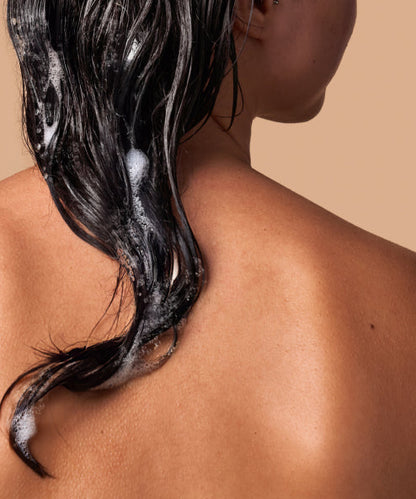 A model facing backwards showing her dark hair lathered in bubbles.